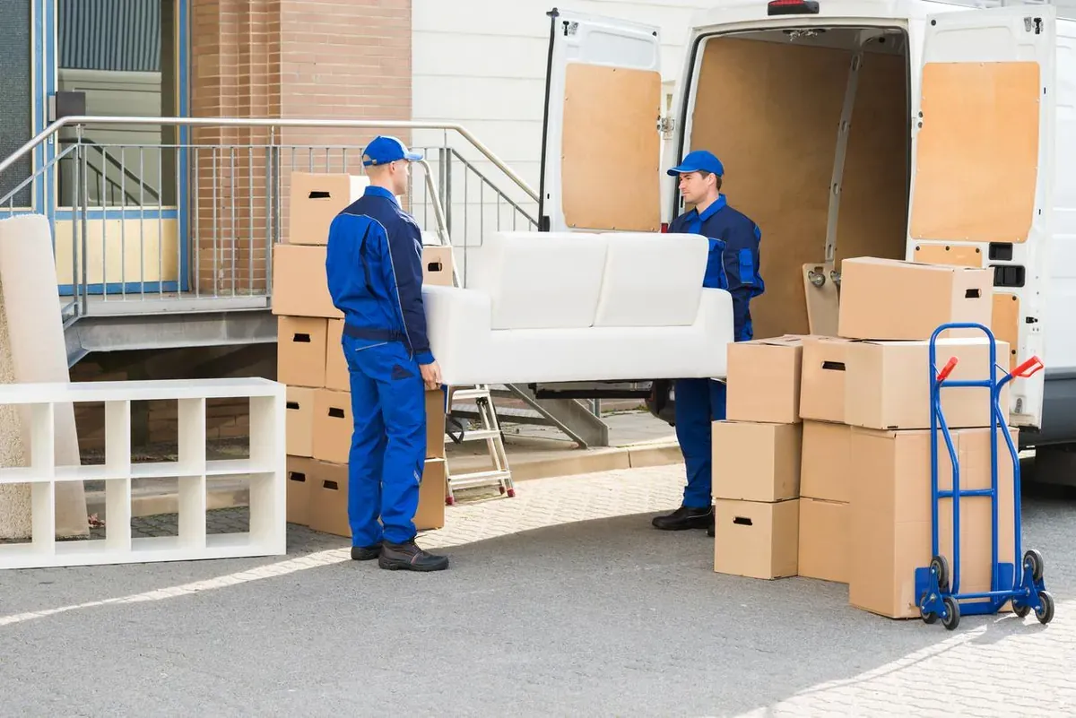 Two movers in blue uniforms unload a white sofa from a moving van.
